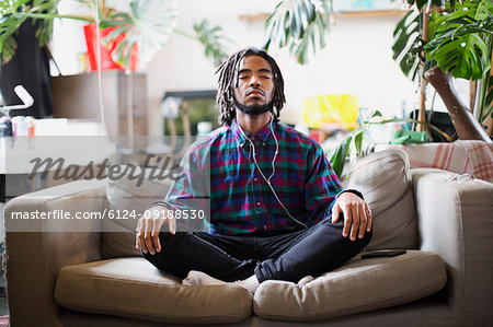 Serene young man meditating with headphones on apartment sofa