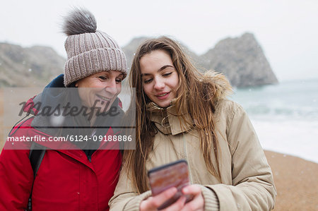 Mother and daughter in warm clothing using smart phone on snowy winter beach