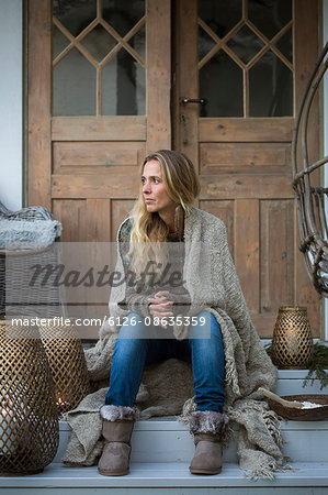 Sweden, Blonde woman wrapped in plaid sitting on house porch