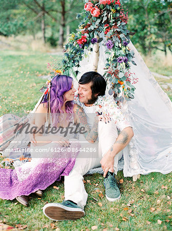 Sweden, Bride and groom sitting on grass by white tent at hippie wedding
