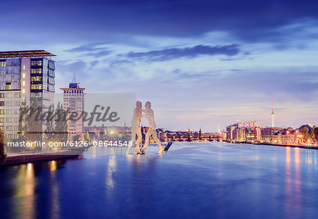 Germany, Berlin, Molecule Man Sculpture on Spree and illuminated riverfront at dusk