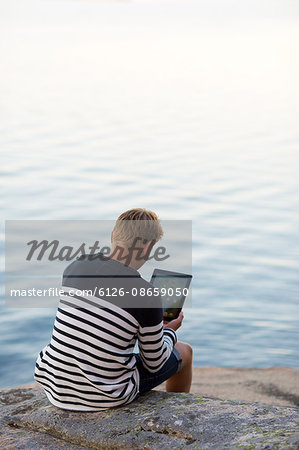 Sweden, Ostergotland, Teenage boy (16-17) sitting on rock and using digital tablet