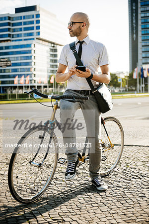 Germany, Berlin, Man with smartphone looking away while leaning on bicycle