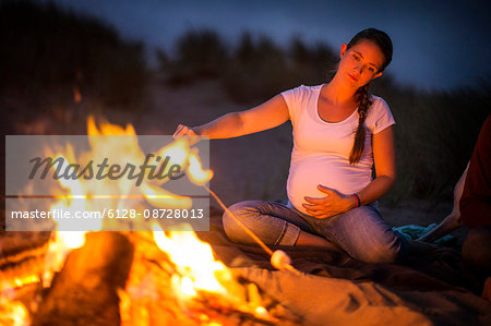 Pregnant young woman enjoys toasting marshmallows over a beach bonfire at night.