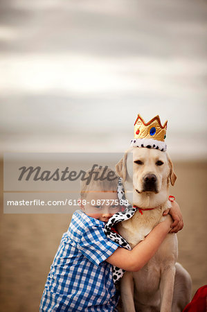 Dog wearing a cape and crown at the beach and being hugged by a little boy.