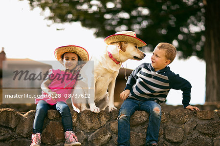 Young boy and girl sitting on a wall with a dog wearing a cowboy hat.