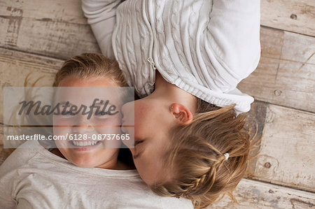 Portrait of a girl being kissed on the cheek by her younger sister while lying on a wooden floor.