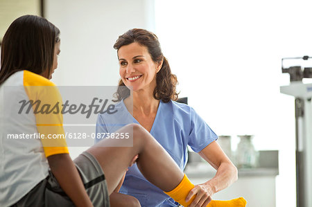 Smiling female doctor holding the injured leg of a teenage girl while sitting together inside a doctor's office.