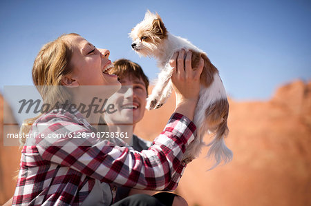 Laughing young woman holding a small dog aloft while sitting next to her boyfriend in a desert.