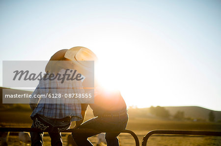 Farmer and his wife sitting on a fence at their ranch and kissing at sunset.