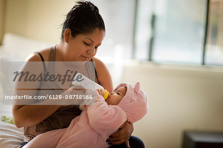 Young mother sitting on the bed giving her baby daughter a bottle.