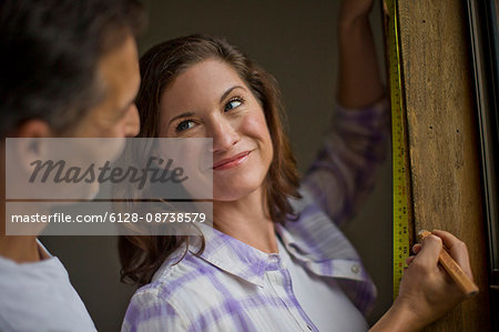Young woman takes a break from measuring the window sill to smile lovingly at her handsome husband who has just come up behind her.
