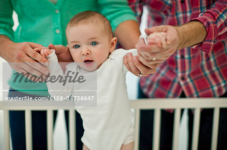 Cute smiling baby boy is supported by his parent's hands as they help him stand in his cot for the first time.