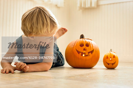 Young boy lies front down on a floor and looks back at a big Jack O'Lantern and a small Jack O'Lantern lit with candles inside next to him as he poses for a portrait.