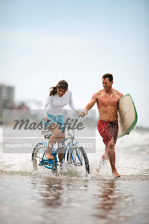 Man with surfboard running alongside a woman on a bike at the beach.