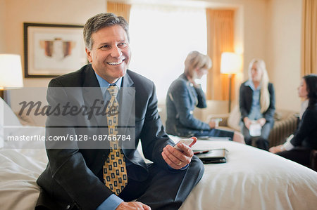 Businessman smiles for a portrait as he sits on the edge of a bed holding his cell phone in a hotel room during on a business trip while his three female colleagues work together in the background.
