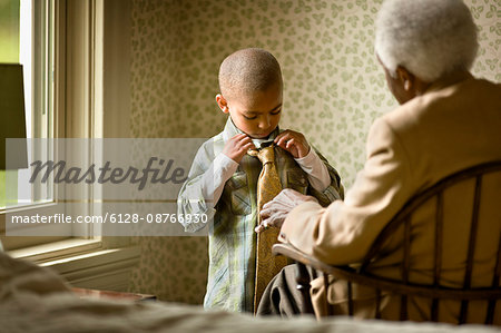 Grandmother sits in a chair and watches her grandson tying a necktie.