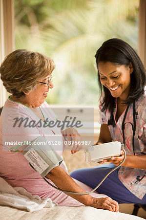 Female nurse uses a blood pressure cuff and blood pressure monitor on a senior woman who sits on a bed.