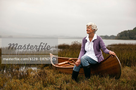 Mature woman smiles and looks away as she poses for a portrait while sitting on the gunwale of a wooden canoe pulled up on a lakeshore.
