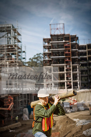 Mid-adult construction worker carrying a large pipe on his shoulder at an outdoor construction site.