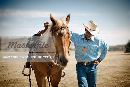 Man wearing a cowboy hat smiles and looks down shyly as he stands next to his saddled horse with one arm raised to rest on the pommel while he poses for a portrait.