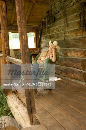 Young Woman Sitting On Rocking Chair On Porch Of Log Cabin