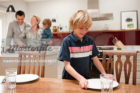 Son helping to lay table with parents and brother visible behind in kitchen