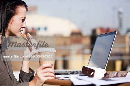 Young woman using a laptop outside while eating a sandwich
