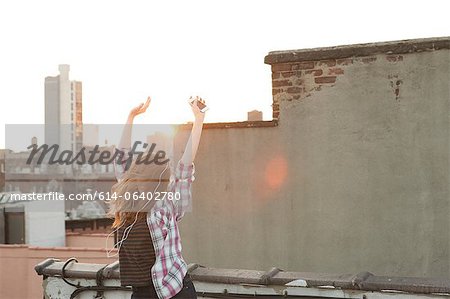 Young woman listening to music with arms raised on city rooftop