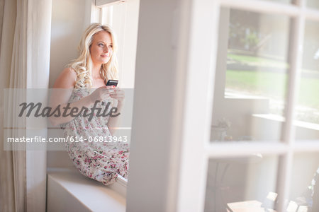 Young woman sitting on windowsill holding smartphone