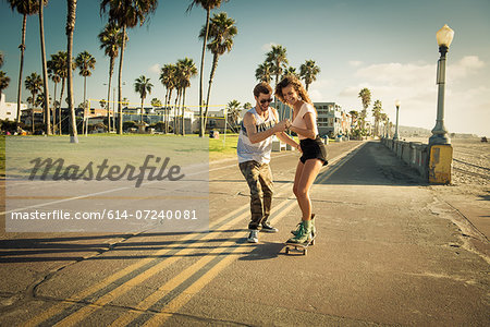 Young woman on skateboard at San Diego beach, boyfriend helping