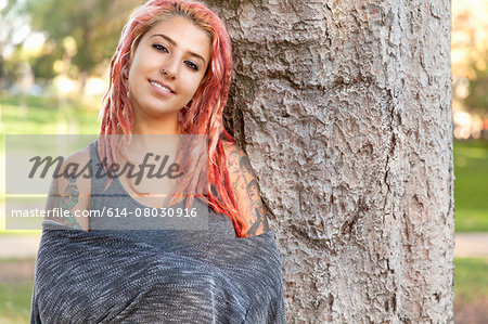 Young woman with pink dreadlocks leaning against tree trunk in park