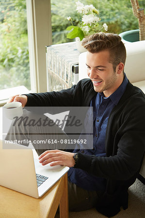 Portrait of smiling man sitting on floor at home looking at laptop and drinking cup of coffee