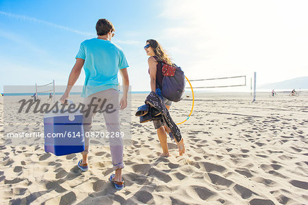 Young couple walking on beach, holding cool box, rear view