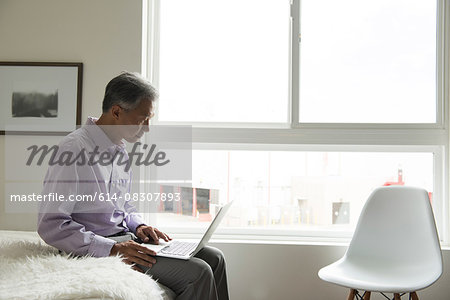 Side view of mature man sitting on edge of bed using laptop