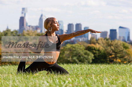 Front view of young woman kneeling on grass stretching leg in yoga position, arm out, looking away, Philadelphia, Pennsylvania, USA