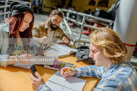 Three adult college students making notes and using smartphones at classroom desk