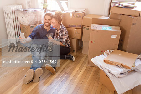 Moving house: Young couple sitting in room full of boxes, holding champagne bottle, taking self portrait with smartphone
