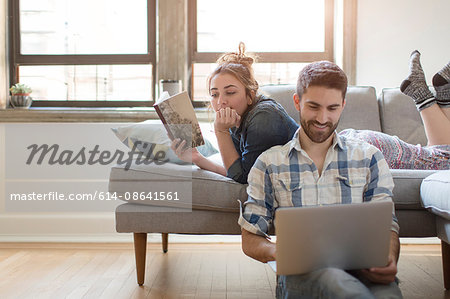 Young couple relaxing at home, young woman reading book, young man using laptop