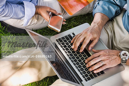 Business man and woman sitting outdoors on grass, using laptop, mid section