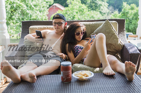 Teenage couple wearing bikini and swim shorts reclining on patio sofa reading smartphones