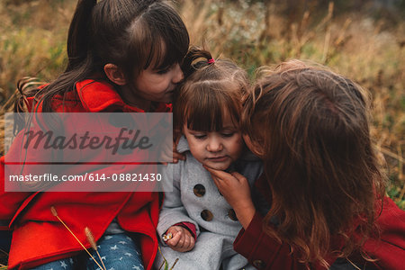 Two girls kissing toddler sister in field