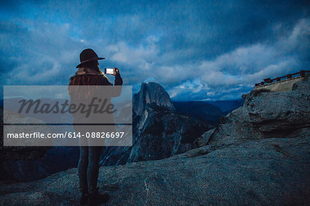 Young woman taking photograph on rock overlooking Yosemite National Park at dusk, California, USA