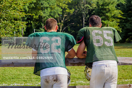 Rear view of two teenage male American football players leaning on playing field fence