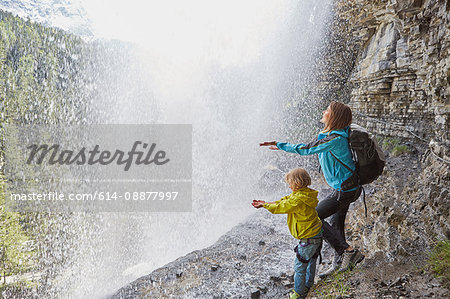 Mother and son, standing underneath waterfall, hands out to feel the water, rear view