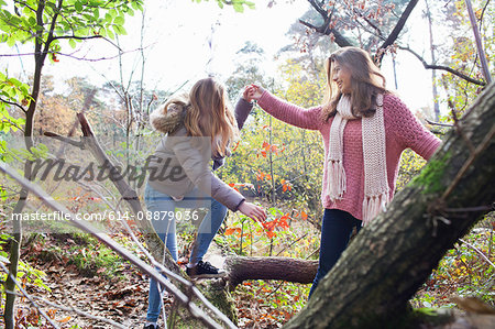 Teenage girl in forest helping frind to climb on fallen tree trunk smiling