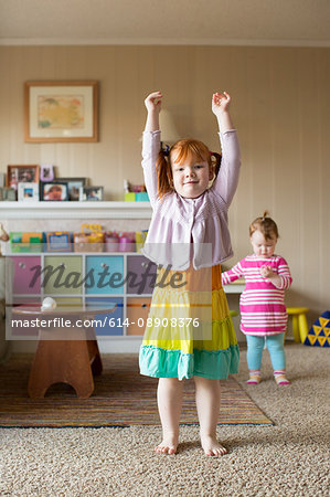 Portrait of  young girl with red hair, arms raised, younger sister walking in background