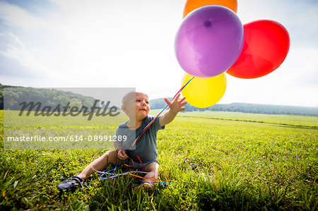 Baby boy sitting in rural field with bunch of colourful balloons