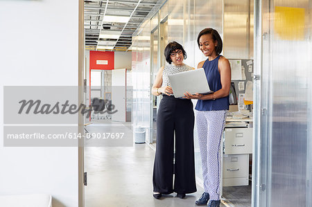 Two women standing in office corridor looking at laptop screen