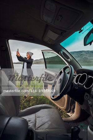 Young woman standing beside Dillon Reservoir, holding smartphone, view through parked car, Silverthorne, Colorado, USA